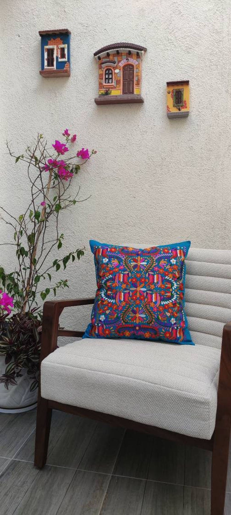 May include: A white chair with a teal blue pillow featuring a colorful floral embroidery pattern. The chair is in front of a white wall with three small decorative houses.