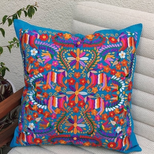 May include: A turquoise blue throw pillow with a colorful embroidered floral pattern. The design features a symmetrical arrangement of flowers, leaves, and swirls in shades of pink, orange, yellow, purple, and green.