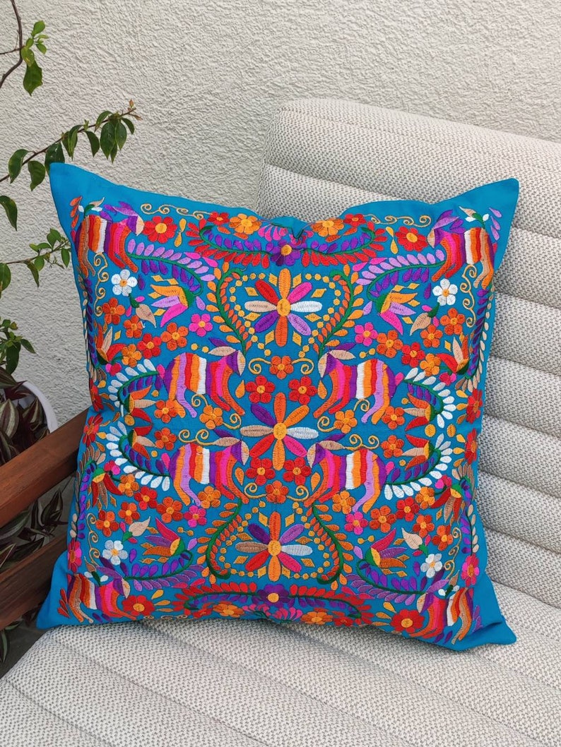 May include: A turquoise blue throw pillow with a colorful embroidered floral pattern. The design features a symmetrical arrangement of flowers, leaves, and swirls in shades of pink, orange, yellow, purple, and green.