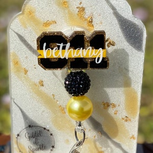 May include: A black and gold badge holder with the number "31" and the name "bethany" written in gold glitter. The holder has a black and gold bead charm and a clear retractable badge clip.