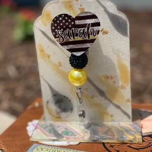 May include: A black and white heart-shaped badge with a thin yellow line and the name "sarah" written in gold glitter. The badge is attached to a black and yellow beaded retractable badge reel.