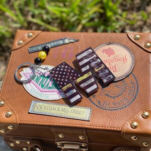 May include: A brown faux leather suitcase with gold accents and various decorative stickers. The stickers include a keychain, a rectangular patch, and three rectangular charms with flag designs. A beaded charm is also present.