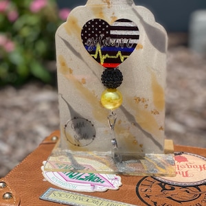 May include: A badge reel with a heart-shaped design featuring the American flag and a blue line, with the word "Gilmore" in cursive. The reel has a black and yellow bead accent and a clear, glittery base.
