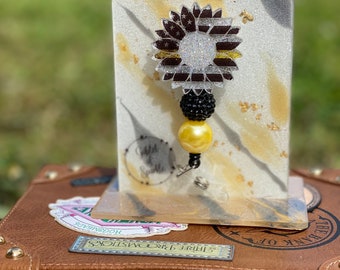 May include: A black and yellow sunflower with a thin yellow stripe across the center is displayed on a white and gold glitter background. The sunflower is attached to a black and yellow beaded charm with a gold spring clip.