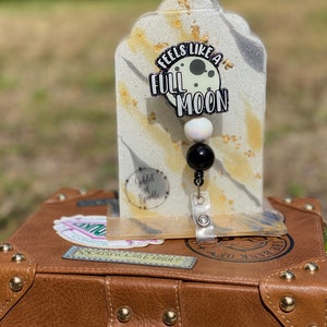 May include: A white and gold resin keychain with a black and white moon charm and the text "Feels Like A Full Moon". The keychain is on a white and gold background with black glitter.