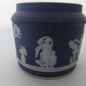 May include: A dark blue ceramic jar with white bas-relief figures and floral designs. The jar has a cylindrical shape with a slightly flared rim and a cream-coloured base. The relief depicts classical scenes.