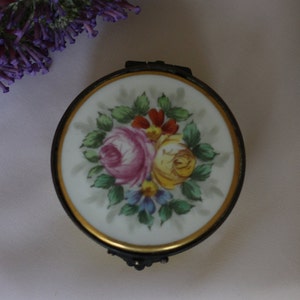 May include: A small, round, porcelain trinket box with a gold rim and floral design. The lid features a hand-painted bouquet of pink, yellow, and red roses with green leaves and blue flowers.