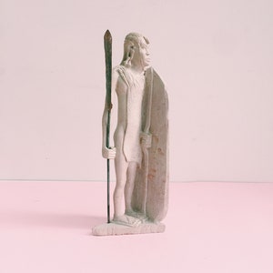 May include: A white stone sculpture of a person holding a spear and a shield. The sculpture is standing on a pink surface.