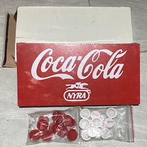 May include: A red Coca-Cola sign with white lettering and the NYRA logo. Two small plastic bags contain red and white bottle caps. A cardboard box is in the background.