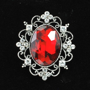 May include: A silver brooch with a large red oval-shaped rhinestone in the center. The rhinestone is surrounded by a silver filigree design with smaller rhinestones.