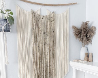 Könnte beinhalten: Eine weiße Wand mit einem Makramee-Wandbehang aus Naturfasern. Der Wandbehang besteht aus drei Abschnitten, wobei der mittlere Abschnitt eine dunklere braune Farbe hat. Es befinden sich zwei Topfpflanzen im Bild, eine mit grünen Blättern und die andere mit einer kleinen grünen Pflanze. Es gibt ein weißes Regal mit einer Vase und anderen Dekorationsartikeln darauf.