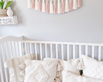 May include: A white crib with a white and pink tassel garland hanging above it. The crib is decorated with white pillows with tassels.