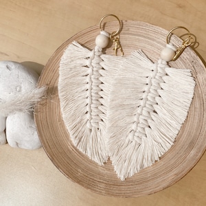 May include: Two white macrame feather keychains with gold rings and wooden beads. The keychains are hanging from a wooden tray.