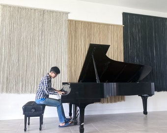 May include: A young person sits at a black grand piano with the lid open, playing the keys. The piano is in a room with three macrame wall hangings, one white, one beige, and one black.