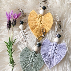 May include: Four handmade macrame keychains in the shape of a heart with fringe. The keychains are in yellow, white, green, and purple. Each keychain has a black lava rock bead and a white wooden bead.