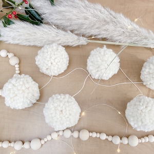 Könnte beinhalten: Eine weiße Pompon-Girlande mit einer Lichterkette und einer Holzperlenkette. Die Girlande hängt an einer hellbraunen Oberfläche.