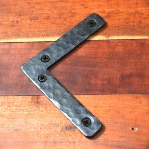 May include: A black metal corner brace with a hammered finish. The brace has four screw holes and is attached to a wooden surface.