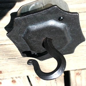 Puede incluir: Un gancho de metal negro forjado a mano con una placa de montaje decorativa en forma de estrella. El gancho se curva hacia abajo y la placa de montaje tiene una superficie texturizada. El gancho está diseñado para colgar objetos.