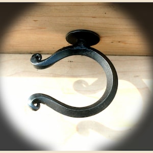 以下が含まれることがあります： 黒色の手鍛造鉄製引き出し取っ手。円形のデザインで、曲がったハンドルの両端に装飾的な渦巻きがあり、円形の取り付けプレートが付いています。 金具は明るい色の木材の表面に取り付けられています。