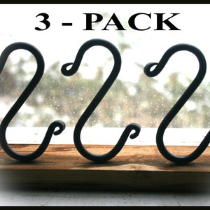 May include: Three black metal S-shaped hooks, labeled "3-PACK", are displayed against a blurry background. The hooks are arranged in a row on a wooden surface.