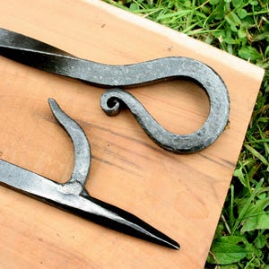 Puede incluir: Dos herramientas de fuego artesanales de metal oscuro. Una herramienta tiene un gancho curvado con un extremo en espiral, y la otra tiene un extremo puntiagudo. Las herramientas se muestran sobre una superficie de madera marrón claro, con hierba verde visible en el fondo.