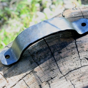 May include: A curved, dark metal handle with two mounting holes. The handle is arched and sits on a weathered, textured wooden surface. The metal has a slightly reflective quality, suggesting a possible coating or finish.