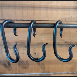 May include: Three black metal S-hooks hanging from a horizontal metal rod. Each hook features a smooth, curved design with a pointed tip at the bottom and a rounded top loop that secures the hook onto the rod, showcasing a dark, matte finish.