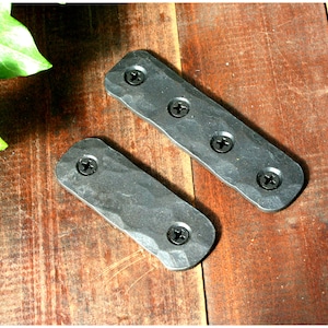 May include: Two black metal rectangular plates with four screw holes each. The plates have a hammered texture and are lying on a wooden surface.