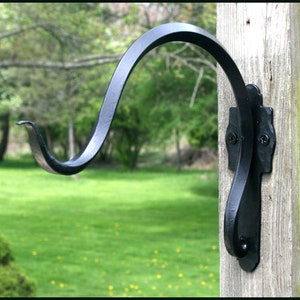 Pode incluir: Gancho preto de ferro forjado para pendurar plantas. O gancho tem um design curvo com uma placa traseira decorativa, montado num poste de madeira. O gancho foi concebido para pendurar cestos suspensos ou outra decoração exterior.