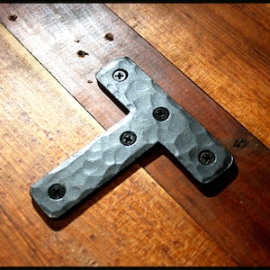 May include: Black metal T-shaped bracket with a hammered finish and five visible screws. The bracket is mounted on a wooden surface.