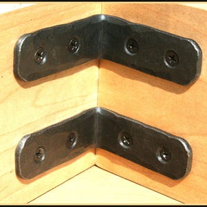 May include: Two black metal corner brackets are attached to a light wood surface. The brackets are rectangular with rounded edges and have four screw holes.