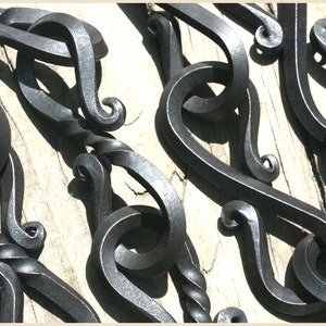 May include: A close-up of several hand-forged metal hooks with decorative swirls. The hooks are black and have a shiny finish. They are arranged in a random pattern on a wooden surface.