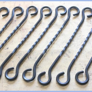 May include: A collection of black metal skewers with a twisted design on the shaft and a looped handle. The skewers are arranged in a row, angled slightly to the right, against a light-colored wooden surface.