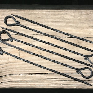 Puede incluir: Juego de seis brochetas de metal negro con un diseño de eje retorcido y asas en forma de lazo. Las brochetas están dispuestas diagonalmente sobre una superficie de madera envejecida, mostrando sus puntas afiladas y asas decorativas. Ideal para asar a la parrilla.