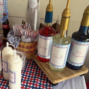 Op de afbeelding: Een soda bar opstelling met drie glazen siroopflessen met gouden pompen gelabeld Cherry, Vanilla en Blue Raspberry, twee witte knijpflessen, een bord met de tekst Star-Spangled Soda Bar, glazen flessen Dr Pepper en Coca-Cola en rietjes.