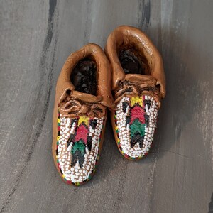 May include: A pair of miniature moccasins. The shoes are brown with intricate beadwork in red, green, yellow, and white. The beadwork forms a geometric pattern on the toe of each shoe. The moccasins are displayed on a gray surface.