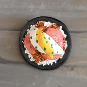 Op de afbeelding: Een miniatuur, handgemaakt ontbijtgerecht. Het zwarte bord bevat een gebakken ei met groene stippen, roze vlees en witte rijst. Het eten heeft een grillig, artistiek uiterlijk.