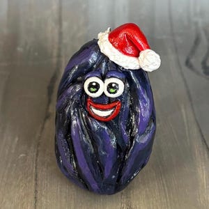 May include: A whimsical, purple, oval-shaped ornament with a smiling face and green eyes, wearing a small red Santa hat with white trim. The ornament has a textured surface and a cheerful expression, perfect for holiday decor.