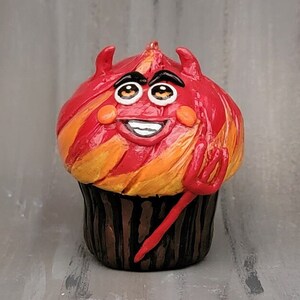 May include: A whimsical cupcake figurine with a devilish design. The red and orange frosted top features a smiling face with horns and a small red pitchfork. The cupcake sits in a brown, striped wrapper.