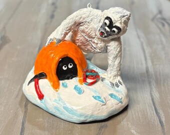 Puede incluir: Adorno pintado a mano que representa un yeti blanco con cara negra y boca roja, inclinado sobre un refugio naranja. Dentro del refugio hay una figura negra con ojos blancos. También hay un piolet rojo y una cuerda.
