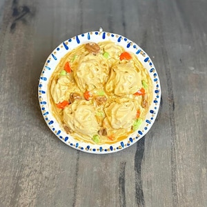 May include: A ceramic ornament showing a bowl of dumplings in a creamy sauce, garnished with vegetables and nuts. The white bowl has a blue and gold dotted rim. The ornament is on a grey surface.