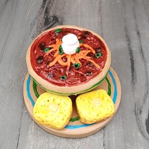 May include: A miniature clay model of a bowl of chili with a dollop of sour cream and two pieces of cornbread on a plate.