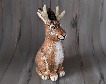 May include: A ceramic jackalope sculpture, a mythical rabbit-like creature with antlers. The jackalope is brown with white details. It is set against a wooden backdrop.