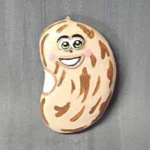 May include: A whimsical, bean-shaped decorative item with a smiling face. The bean is painted in a light beige color with brown markings. It has a white mouth, pink cheeks, and green eyes. The item is likely a magnet or a decorative piece.