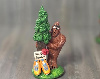 Puede incluir: Una figura de criatura marrón y peluda de pie junto a un pino verde sobre una base verde. La criatura tiene ojos grandes y la boca abierta. Un letrero blanco con el texto "FROM SANTA" está sujeto al árbol, con dos zapatos naranjas debajo.