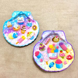 May include: Two decorative scallop shells painted with pastel watercolour backgrounds and adorned with miniature resin charms of various desserts, including cupcakes, ice cream cones, popsicles, and doughnuts. One shell features a central cupcake with the number 30.