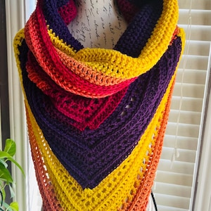 May include: A handmade crochet shawl in a vibrant gradient of colors, including yellow, orange, red, and purple. The triangular shawl is displayed on a mannequin, showcasing its intricate stitch pattern and cozy design. This accessory is perfect for adding a pop of color to any outfit.