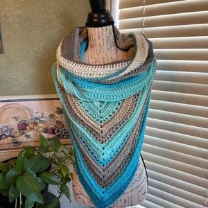 May include: A handmade crochet shawl in a triangular shape, displayed on a mannequin. The shawl features a gradient of colors, including shades of blue, gray, and cream. The texture of the crochet is visible, showcasing the intricate stitch work. The shawl is designed to be worn around the neck and shoulders.