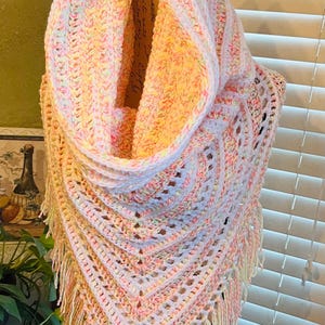 May include: A handmade, pastel-colored crochet shawl with a cowl neck and fringe. The shawl features a mix of white, pink, yellow, and green yarn, creating a soft, textured look. The openwork design and fringe add a touch of elegance to this cozy accessory.