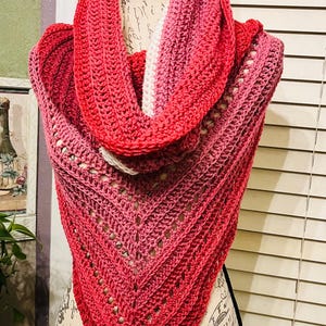 May include: A handmade, crochet shawl in shades of red and pink, displayed on a mannequin. The shawl features a gradient color pattern and a triangular shape. The openwork design adds a delicate touch to this cozy accessory. This item is perfect for adding warmth and style.
